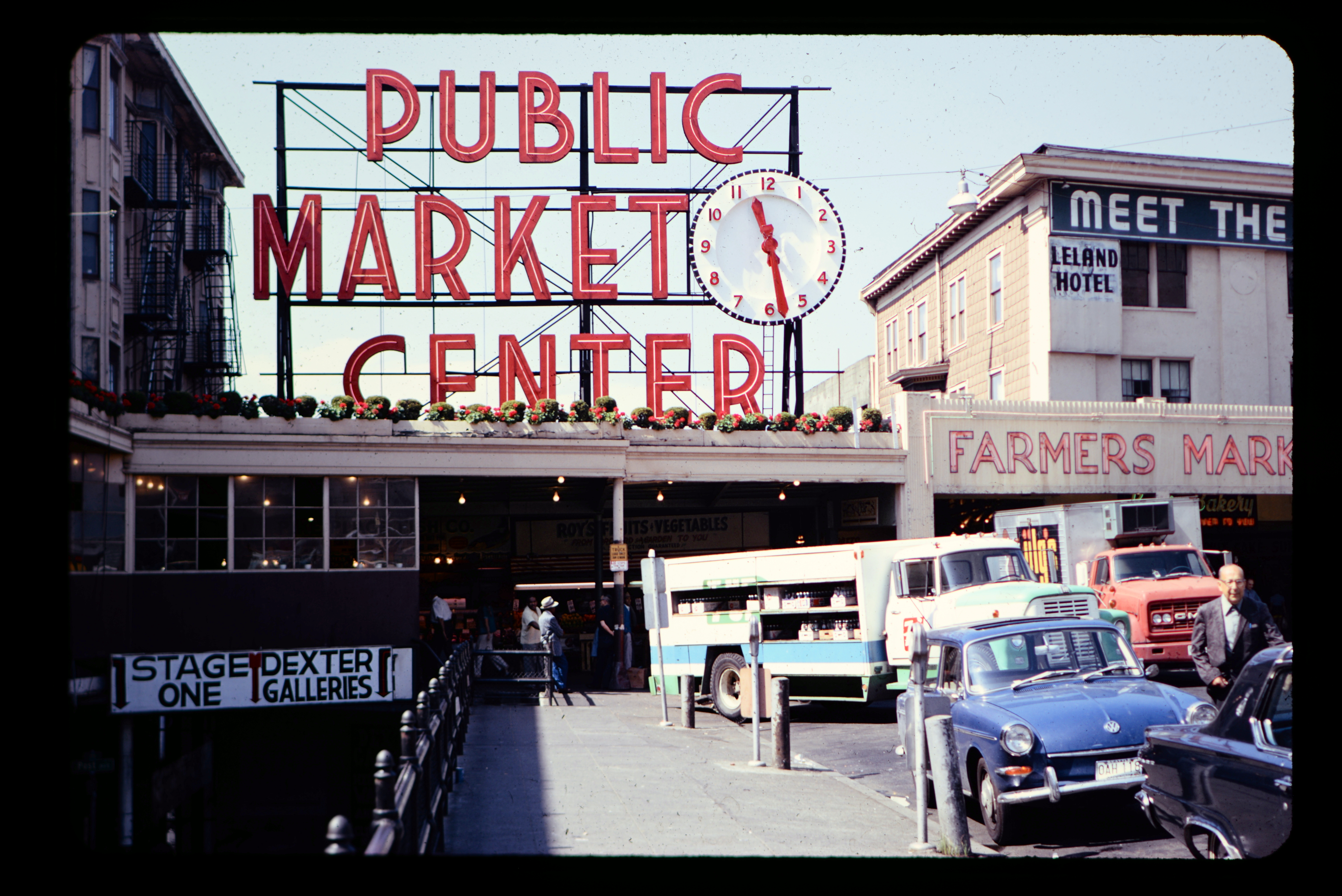 A sign reading "Public Market Center" with a clock atop a building  housing a farmer's market.  Cars and trucks from the 1970s are parked in front of the building.