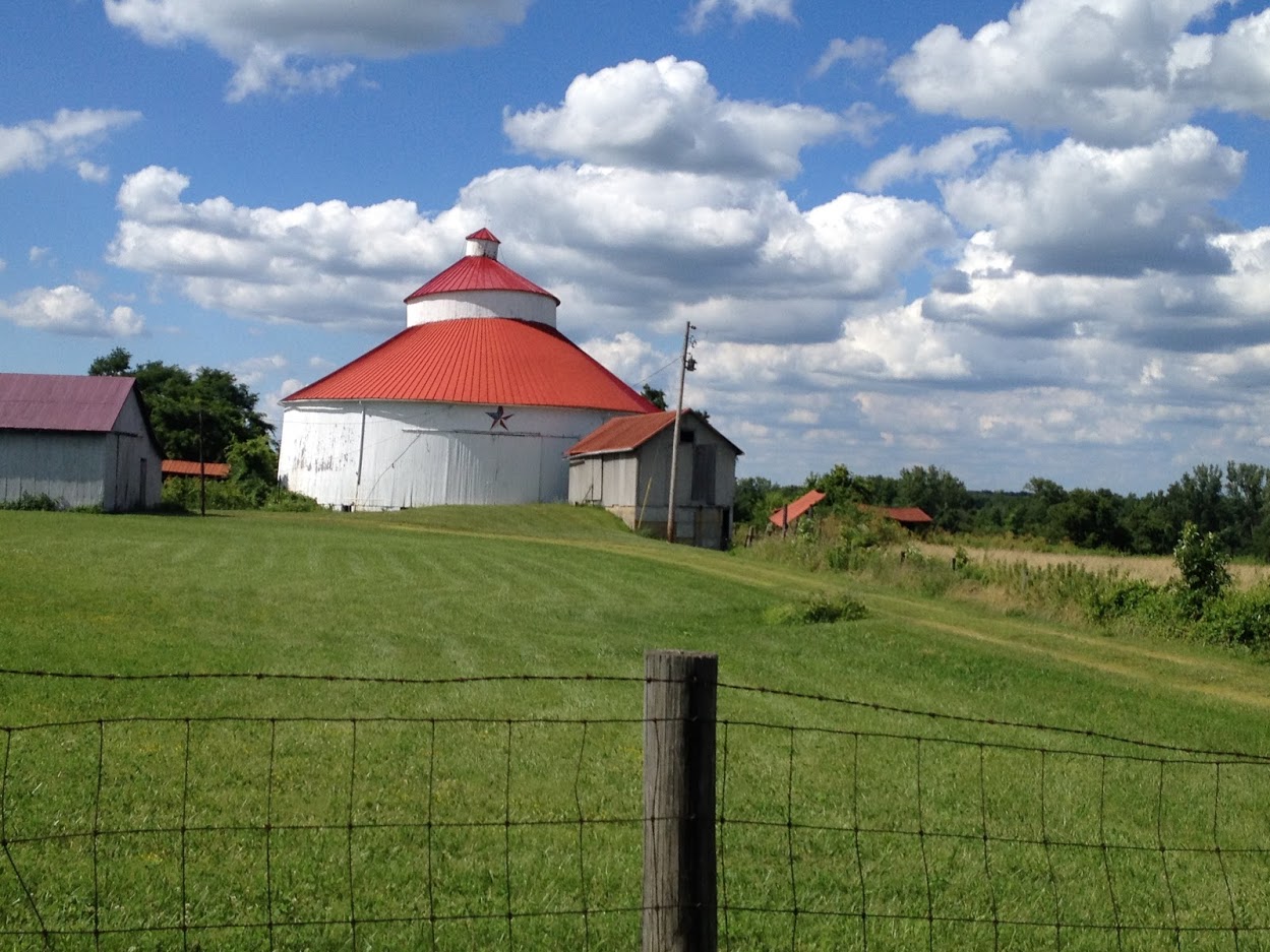 A photo of round, white barn with a red roof in a field. Puffy clouds float in the sky. 
