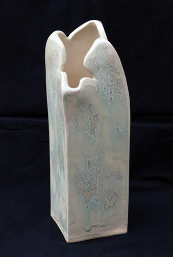 An extruded ceramic vase sits at the center of this image. It is based on a square shape which is three times as tall as it is wide. The top of the form has a scalloped edge which resolves by bending and caving in slightly. On the exterior of the piece, crystals in the fired glaze have produced an irregular pattern of silver lined shapes against a pale green background. The interior is off-white.