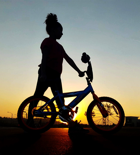 A photograph at sunset that highlights the silhouette of a child on a bicycle, strongly contrasted by the twilight sky.