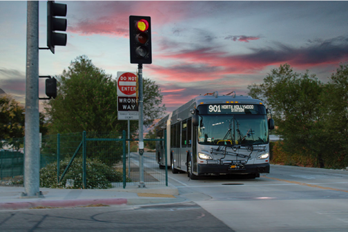 A sunset image of a 901 North Hollywood line Metro bus sits at an intersection, waiting for the red light to change. Surrounding the bus are trees, the light post in center frame with a “Do Not Enter” sign behind it and the adjacent light and sidewalk. 