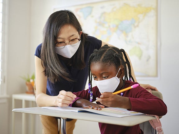An elementary aged school student and teacher in a classroom wearing a mask for protection against infectious disease. Image courtesy of RichLegg, iStock
