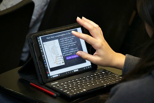 A CSUN student is shown browsing and taking notes on a tablet device during a class lecture.