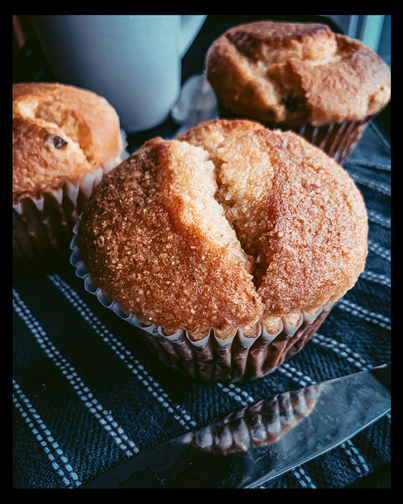 On the commercial photography food project, the image displays three muffins in close detail, resting on a black and white stripped dish cloth. There is a butter knife that spans the foreground of the frame and a soft focus, light grey mug in the background.
