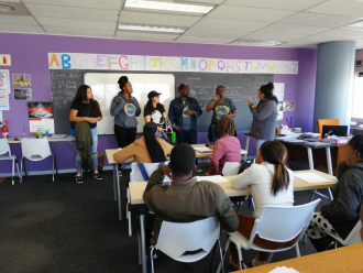 A group of standing people speaking in ASL to a group of seated listeners in a classroom