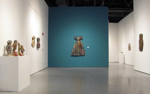 Install image looking into the main Gallery from the front door. Artworks from left to right:  Group of bound dolls with costume jewelry and string.  Two baby dresses covered in costume jewelry.  Large woman’s dress made out of zippers and pins.  Cocktail