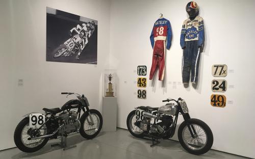  Install shot of the back left room of the Main Gallery.  Two Motorcycles National number 98.  In the corner, a trophy for the rider, Hateley Number 98 who’s leathers are on the right wall.  On the Left wall an image of rider Number 10 in skeleton leather