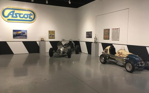  Install shot of front room in Main Gallery.  Walls were painted with black and white stripes to look like a crash wall.  Two midget cars pictures.  Number 49 spike jones. The walls contain original posters from the races, and the original Ascot sign that