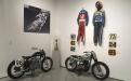  Install shot of the back left room of the Main Gallery.  Two Motorcycles National number 98.  In the corner, a trophy for the rider, Hateley Number 98 who’s leathers are on the right wall.  On the Left wall an image of rider Number 10 in skeleton leather